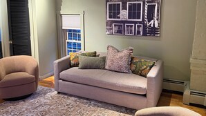 Een woonkamer met een beige bank, twee fauteuils, een kamerbreed tapijt en een aan de muur hangend schilderij van een vintage kledingwinkel.