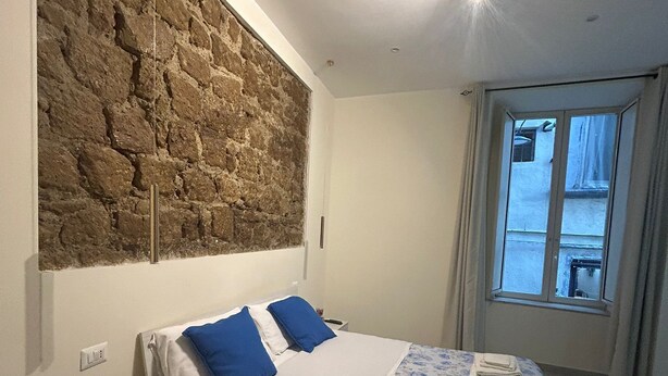 Een moderne slaapkamer met een accentmuur van steen, een bed met blauw-witte beddengoed, een nachtkastje met een plant en een raam met gordijnen.