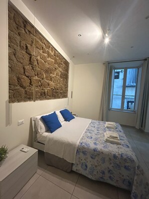 Um quarto moderno com parede de pedra decorativa, cama com roupa de cama azul e branca, mesa de cabeceira com uma planta e janela com cortinas.