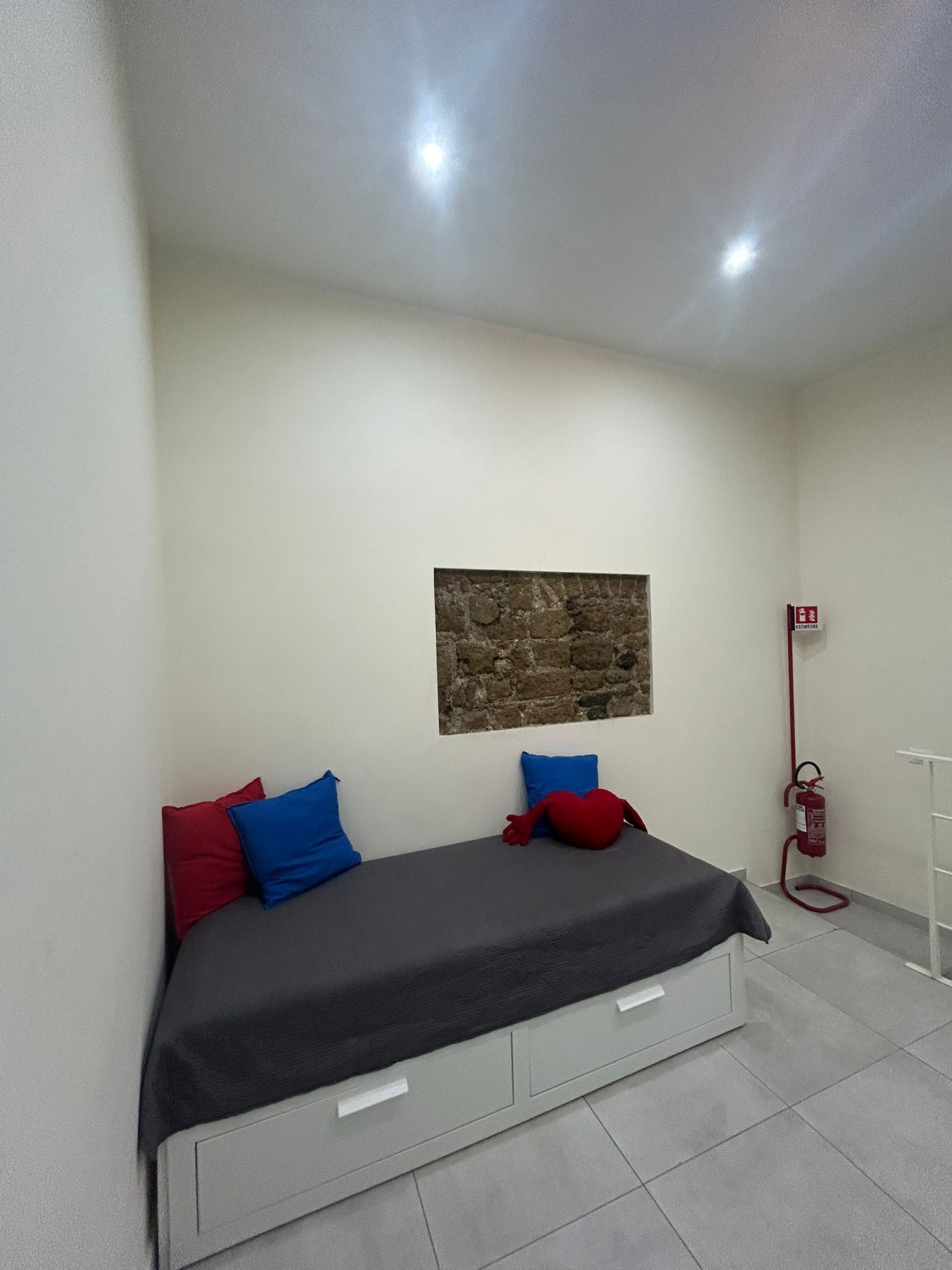 A small, modern bedroom with a bed, a stone accent wall, a fire extinguisher, and a white ceiling.