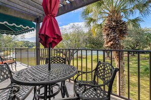 A covered outdoor patio with a round table and chairs, a red umbrella, and a view of a lake and palm trees. - Beautiful Naples Vacation Rental w/ Shared Pools (Naples)