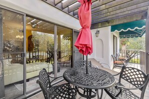 A covered outdoor seating area with a round table and chairs, a red umbrella, and a view of the garden through large glass doors. - Beautiful Naples Vacation Rental w/ Shared Pools (Naples)