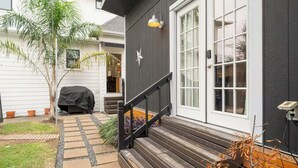 Ein Haus mit dunkler Fassade, einer überdachten Grillgelegenheit, einer Holztreppe und einem kleinen Garten mit Topfpflanzen.
