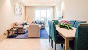 A modern living room with a sofa, a coffee table, and a dining table with chairs. There is a large window providing natural light and a view of the city.