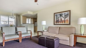 Habitación de hotel con sofá, sillón, reposapiés, un cuadro de palmeras, una lámpara y vistas al exterior.