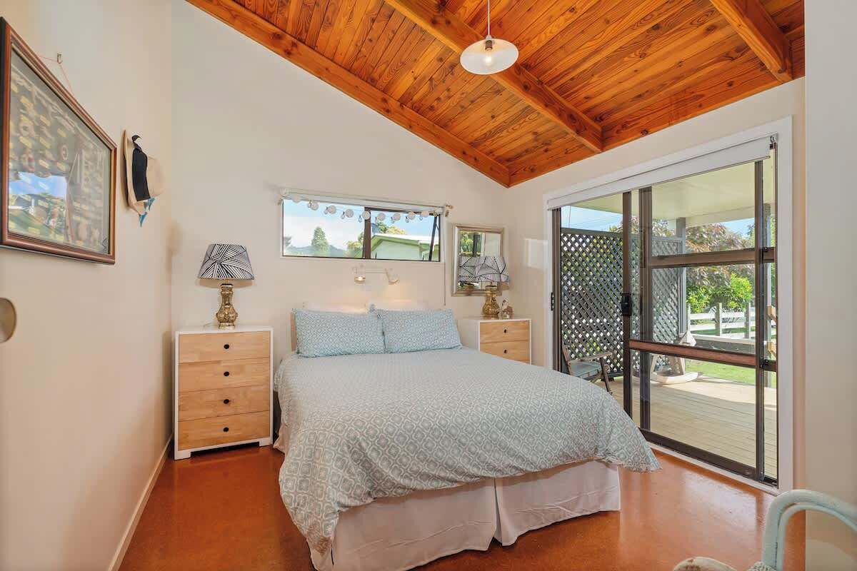 A bedroom with a wooden ceiling, a bed with a patterned bedspread, two bedside tables with lamps, and a large sliding glass door leading to an outdoor area.