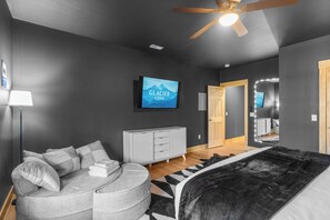 Ein modernes Schlafzimmer mit grauem Sofa, einem schwarz-weißen Teppich, einem großen Bett, einem Fernseher, der 'The Glacier Cove' zeigt, und einer Holztür.