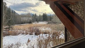 Blick aus einem Fenster mit sichtbarer Holzbalkenstruktur auf eine verschneite Landschaft mit Bäumen und Bergen in der Ferne.