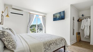 A bedroom with a bed, a window with curtains, a painting on the wall, and a walk-in closet with hanging robes.