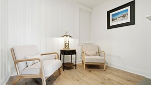 A room with two light-colored armchairs, a small round table with a lamp, and a framed picture on the wall.