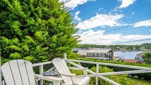 A wooden deck with two white Adirondack chairs and a small table, overlooking a residential area with houses and a body of water.