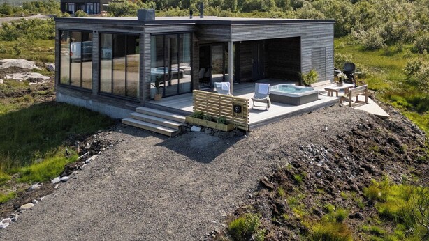 Une maison moderne en verre, dotée d’une terrasse et d’un jacuzzi, entourée de montagnes et de verdure.