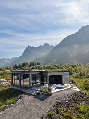 Une maison moderne en verre, dotée d’une terrasse et d’un jacuzzi, entourée de montagnes et de verdure.
