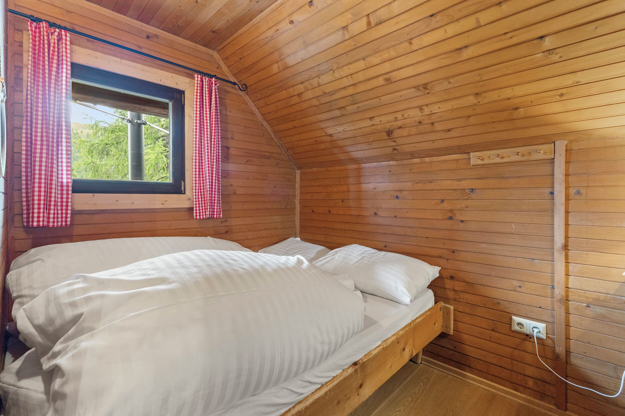 A wooden cabin bedroom with a bed, a window with red and white checkered curtains, and a wall-mounted power outlet.