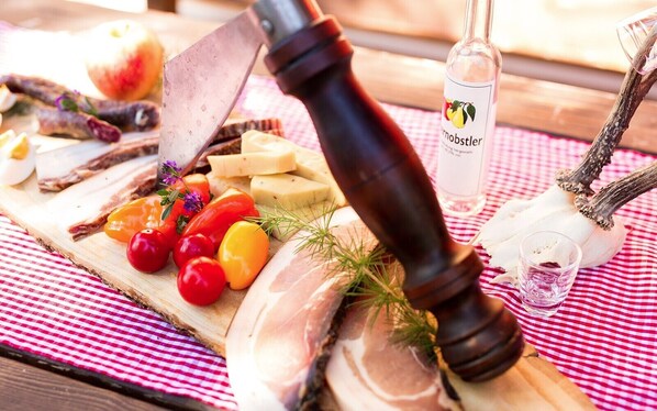 A wooden board with various foods including cheese, tomatoes, and fish, a pepper grinder, and a bottle of drink on a red and white checkered tablecloth.
