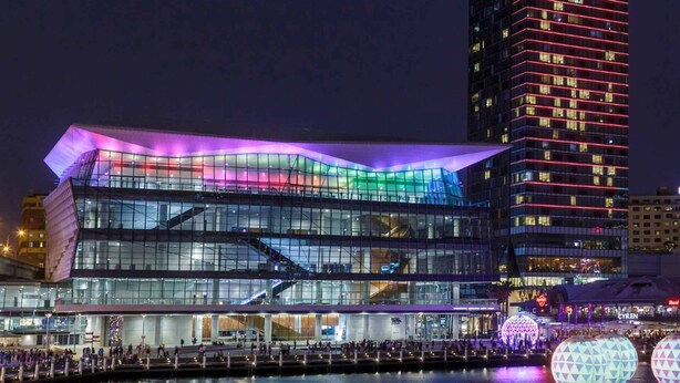 Een modern gebouw met een glazen gevel en een gebogen dak, gelegen naast een waterpartij met verlichte bollen.