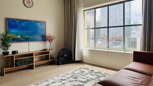 Een moderne woonkamer met een groot raam, een bank, een houten tv-meubel, een schilderij en een ventilator.