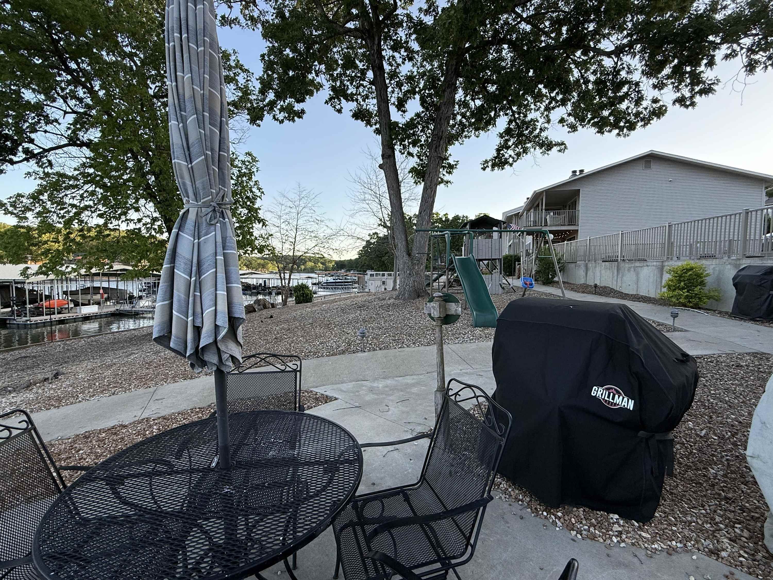 Un patio al aire libre con una mesa y sillas de metal redondas, una parrilla cubierta y un columpio en el fondo.