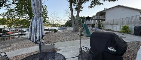 Un patio al aire libre con una mesa y sillas de metal redondas, una parrilla cubierta y un columpio en el fondo.