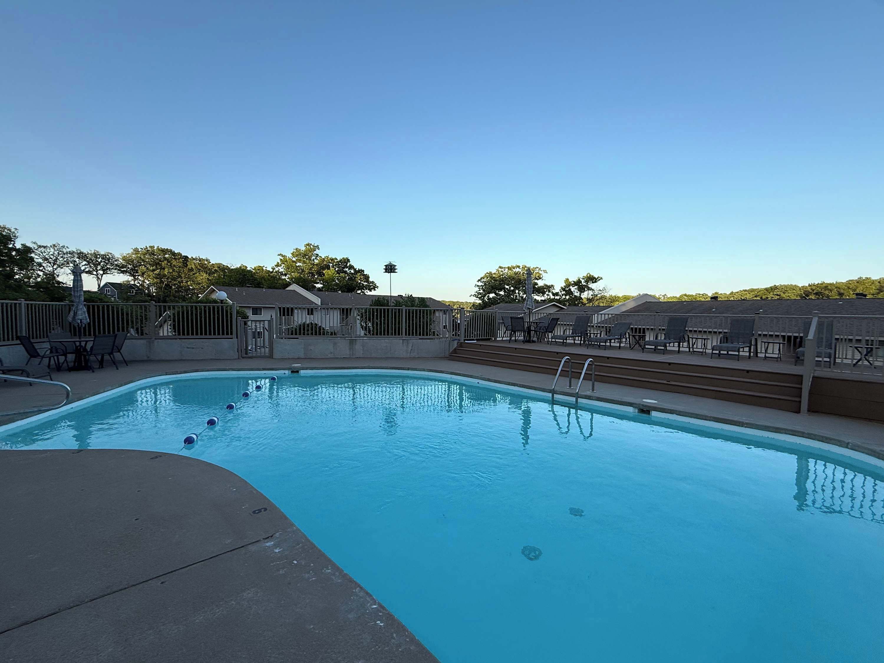 A swimming pool with a clear blue water, surrounded by lounge chairs and umbrellas, and a fence enclosing the area.
