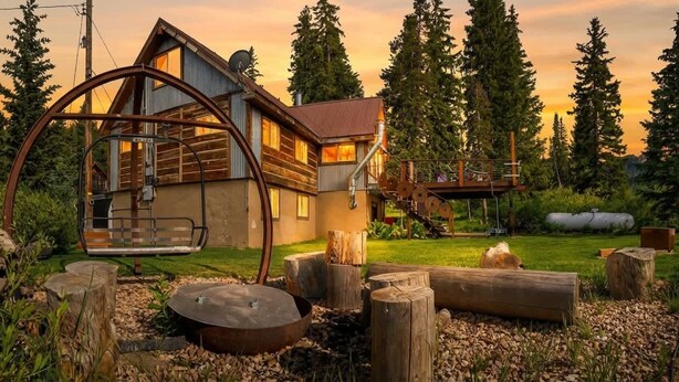 Une cabane rustique dotée d’une roue à eau en métal, d’une terrasse en bois et offrant une vue sur de grands arbres.