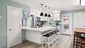 Cocina moderna con armarios blancos, una isla central con taburetes de bar, iluminación colgante y vistas al exterior a través de un ventanal.