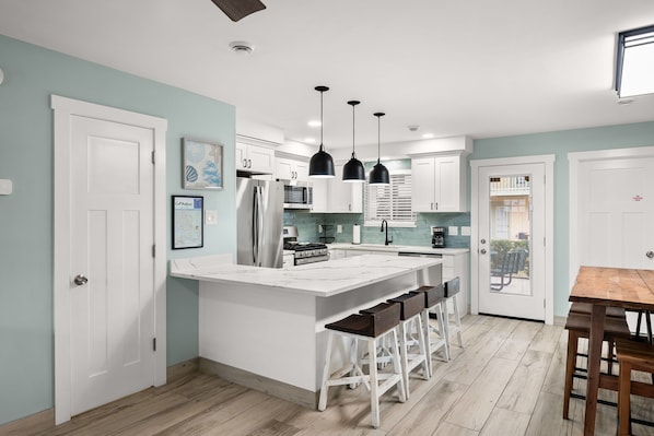 A modern kitchen with white cabinets, a central island with bar stools, pendant lighting, and a view through a glass door to the outdoors. - Beautifully Renovated! W/Golf Cart Available! (Panama City Beach)