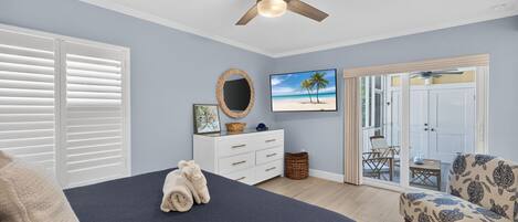 A bedroom with a bed, a ceiling fan, a dresser, a TV, and a view of the outside.