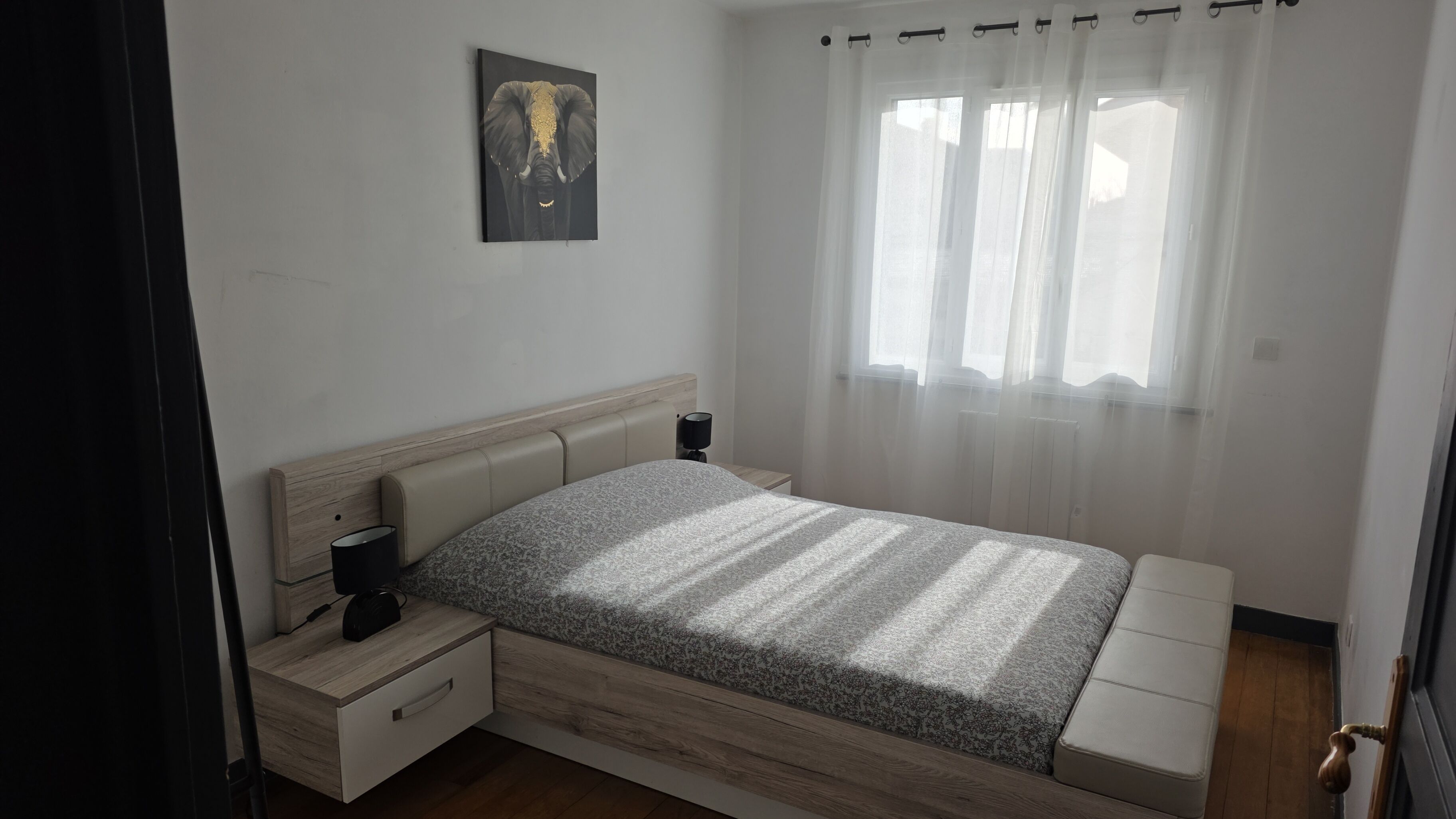 Ein Schlafzimmer mit Bett, Nachttischen, einer Lampe, einem Gemälde und einem Fenster mit durchsichtigen Vorhängen.