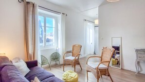 Un salon moderne avec un canapé violet, deux fauteuils en osier, un pouf jaune et une grande fenêtre avec des rideaux.