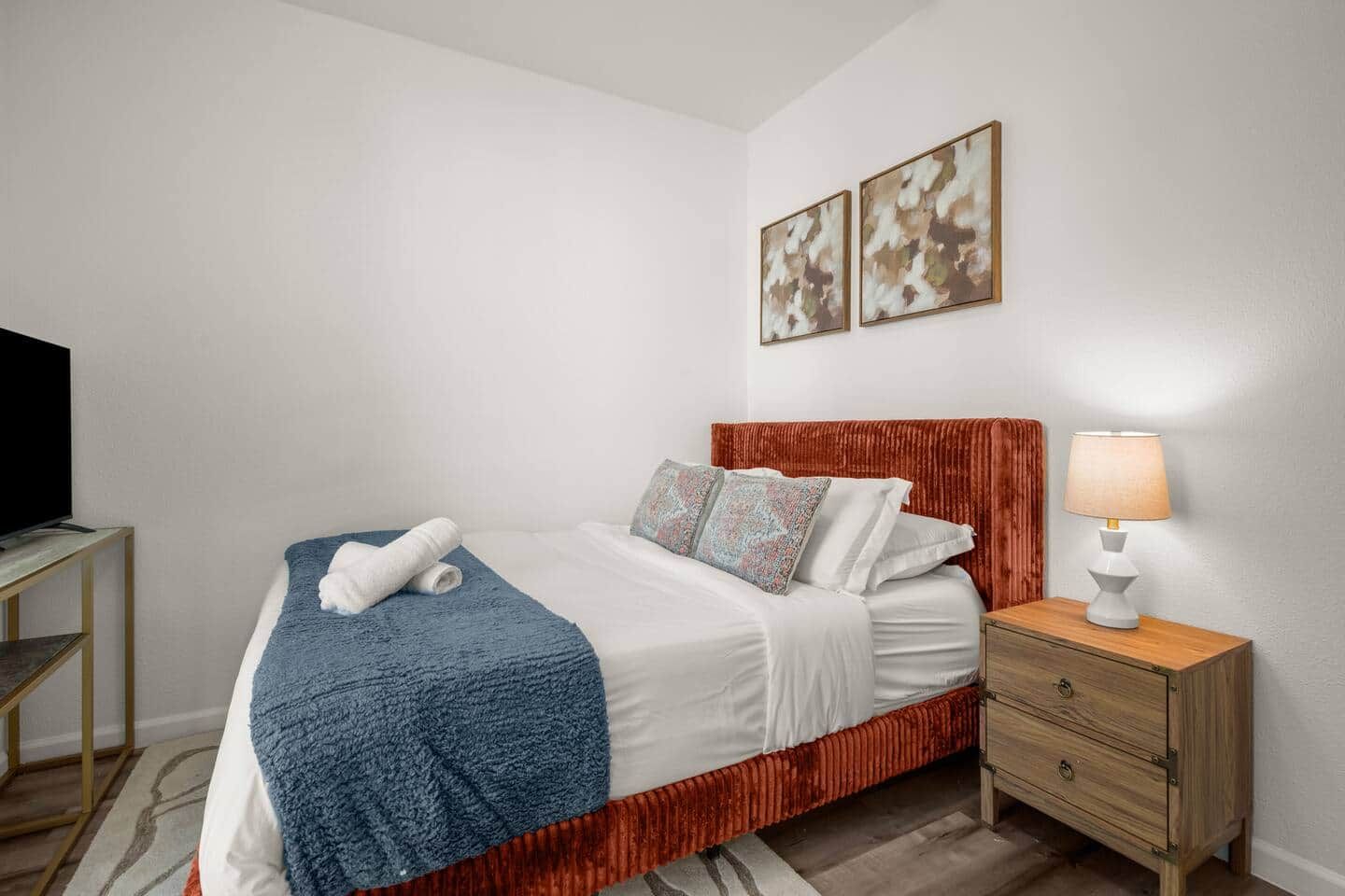 A neatly made bed with a blue blanket, white pillows, and a red headboard. There is a bedside table with a lamp and a TV on a stand.