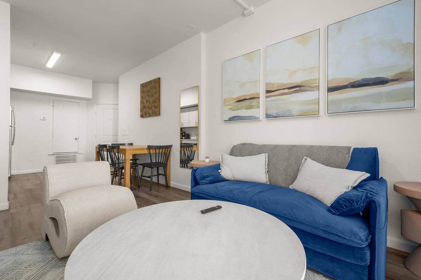 Un salon moderne avec un canapé bleu, deux poufs blancs, un coin repas avec une table et des chaises en bois, et des œuvres d’art abstraites accrochées au mur.