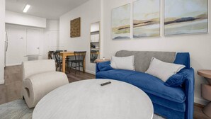 Ett modernt vardagsrum med en blå soffa, två vita sittpuffar, en matplats med ett träbord och stolar samt abstrakt väggkonst.