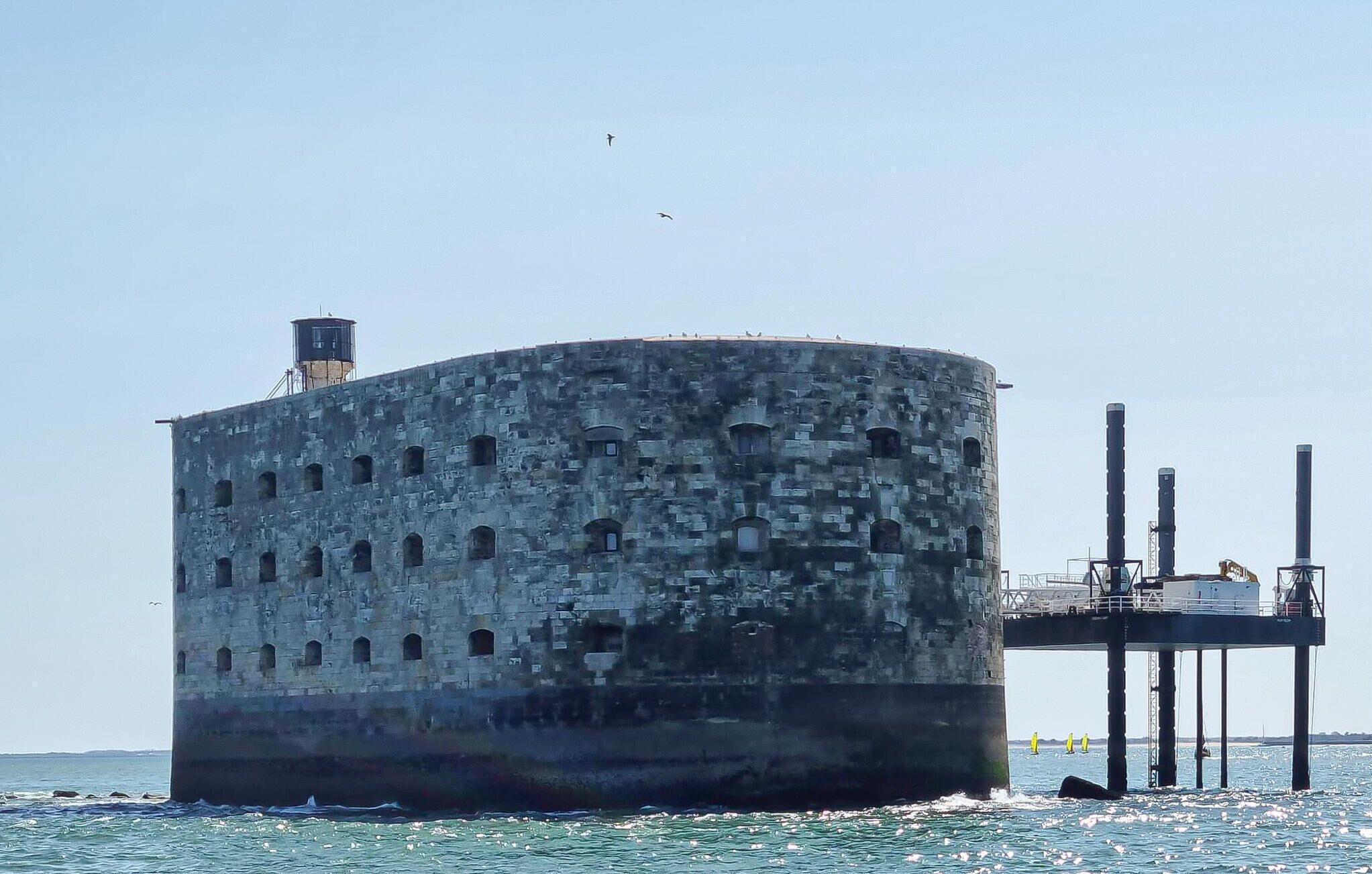 Eine große, zylindrische Steinstruktur mit mehreren Rundbogenöffnungen, einem kleineren Turm obenauf und einem Steg, der ins Wasser ragt.