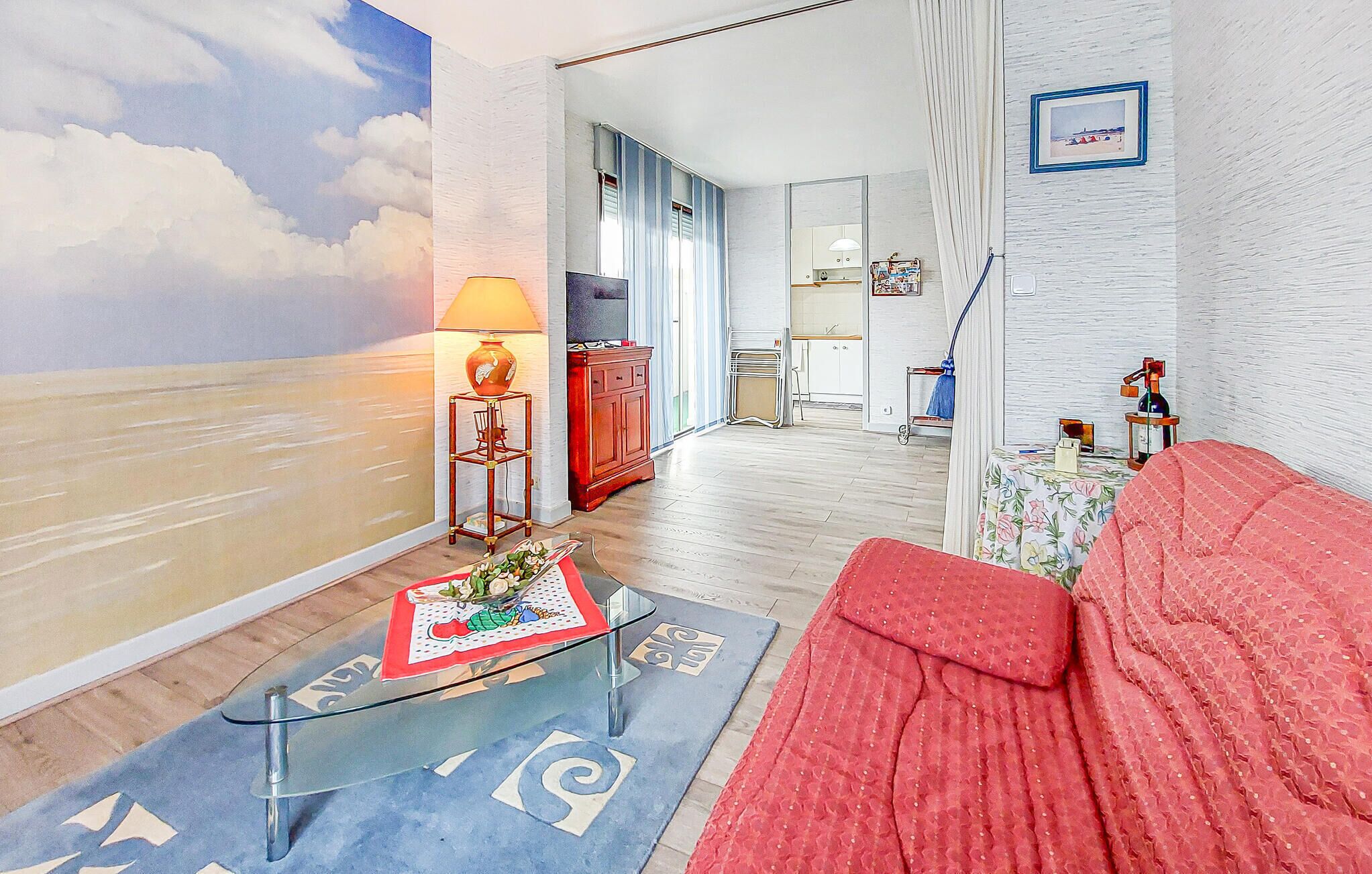 A living room with a red sofa, a glass coffee table, a lamp, and a wall mural of clouds.