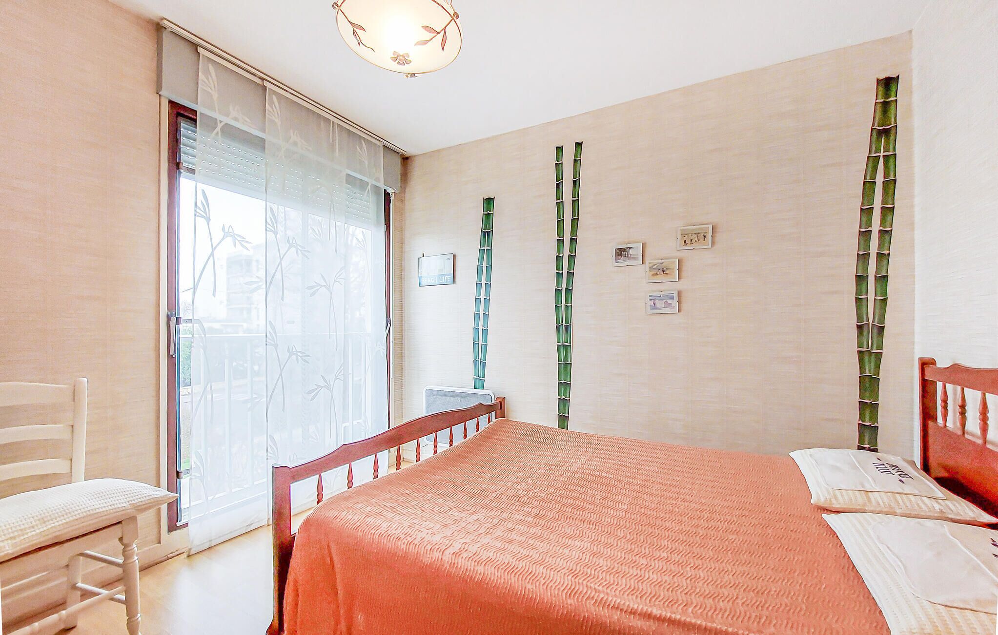 Un dormitorio con cama, una silla, una ventana con detalles de bambú y una pared con cuadros.