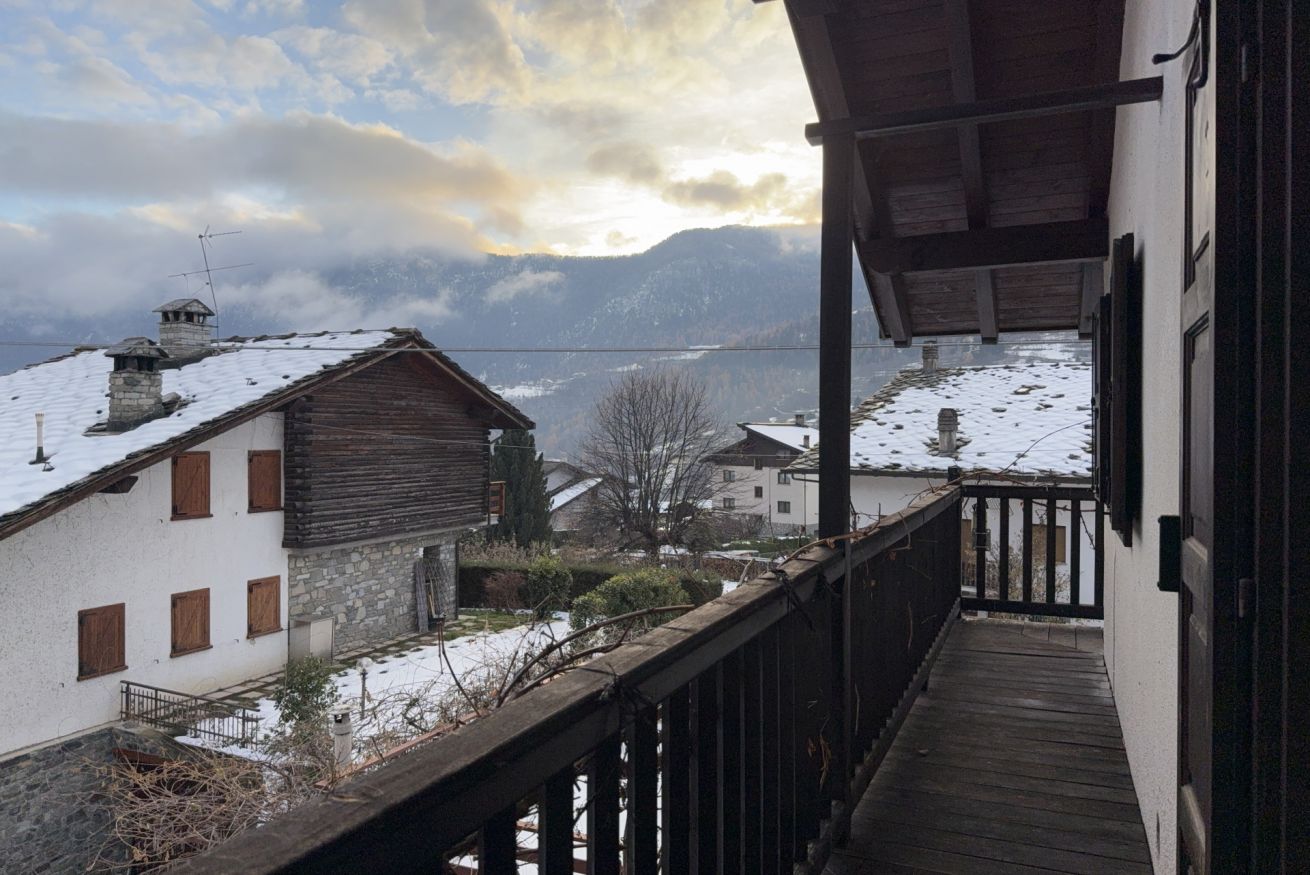 A wooden balcony overlooking a snowy village with stone buildings and a mountain backdrop.