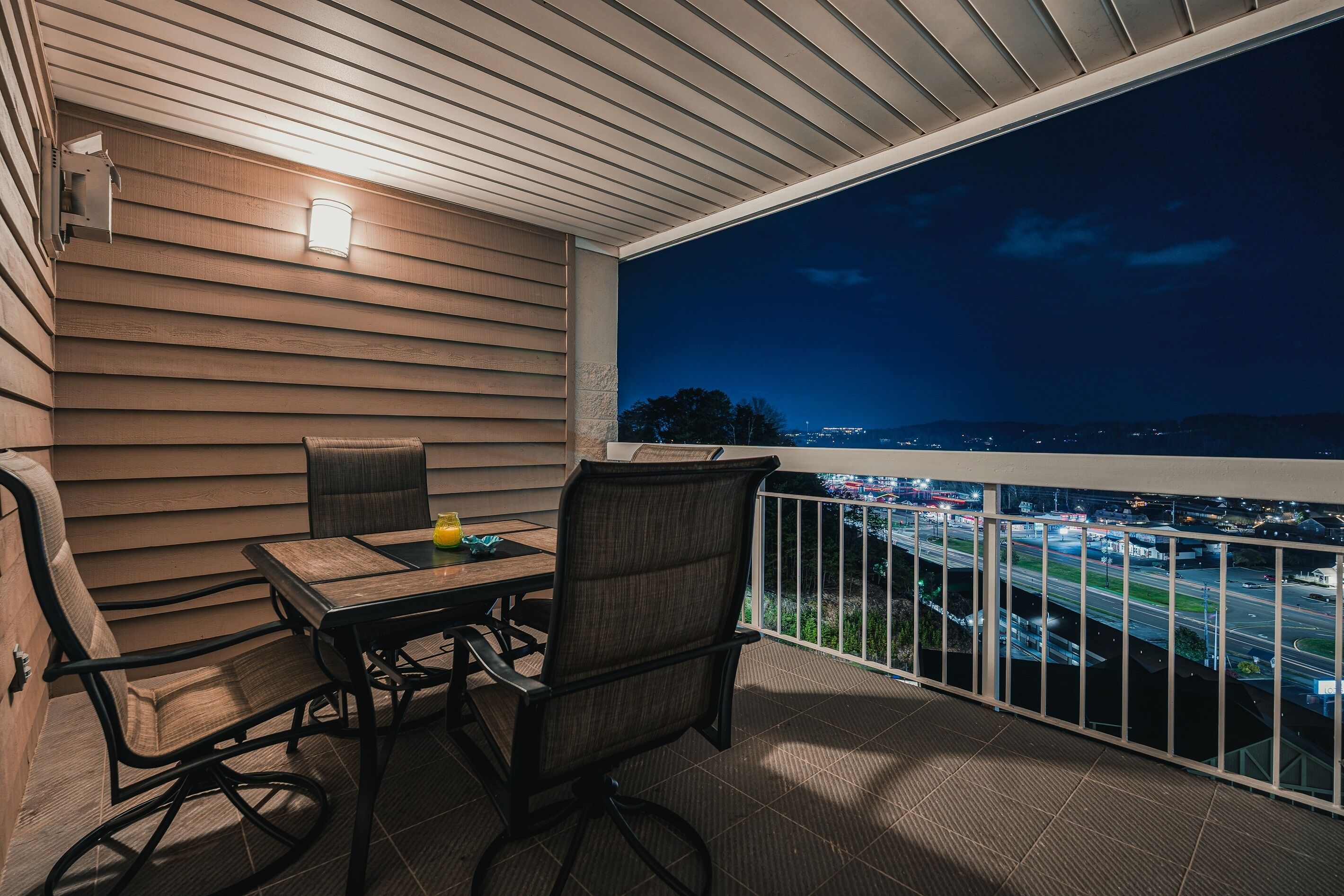 An outdoor patio with a table and chairs, a railing, and a view of a cityscape at night.