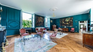 Een ruime kamer met een groot raam, een kroonluchter en een visgraat parketvloer.