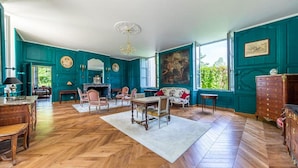 Een ruime kamer met een visgraatmotief houten vloer, teal muren, een open haard en een groot raam met uitzicht op groen.
