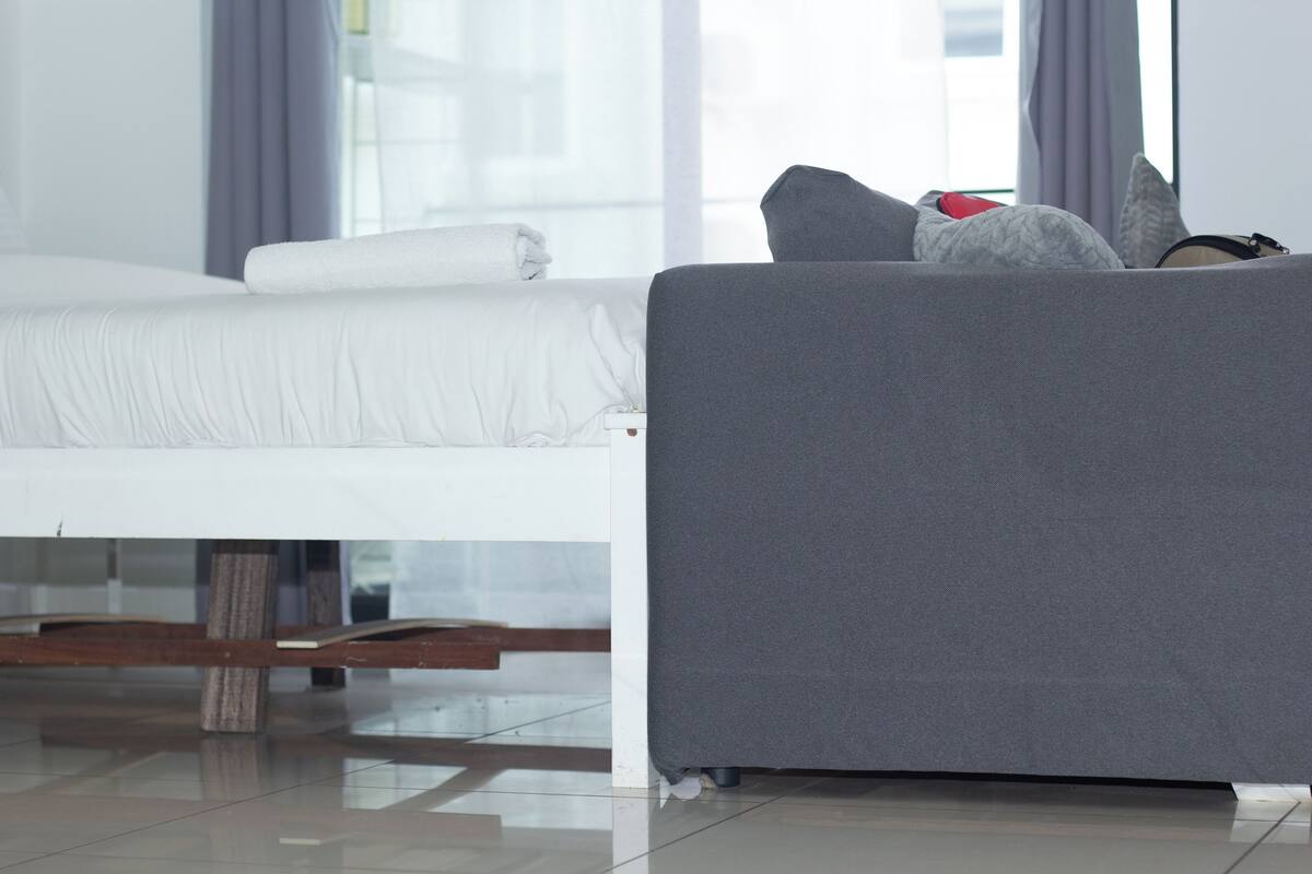 A modern hotel room with a neatly made bed, a grey sofa, and a view of the city through large windows.