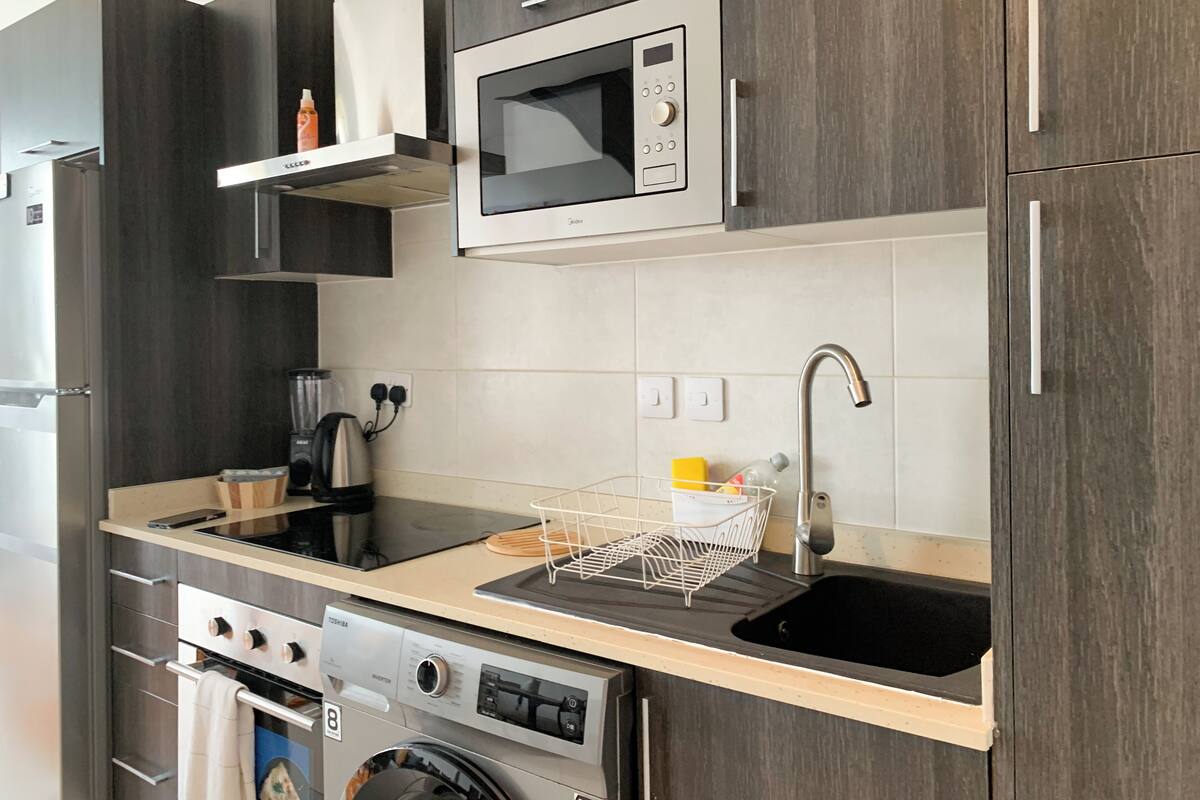 A compact kitchen with a built-in microwave, a stainless steel sink, a kettle, and a washing machine.