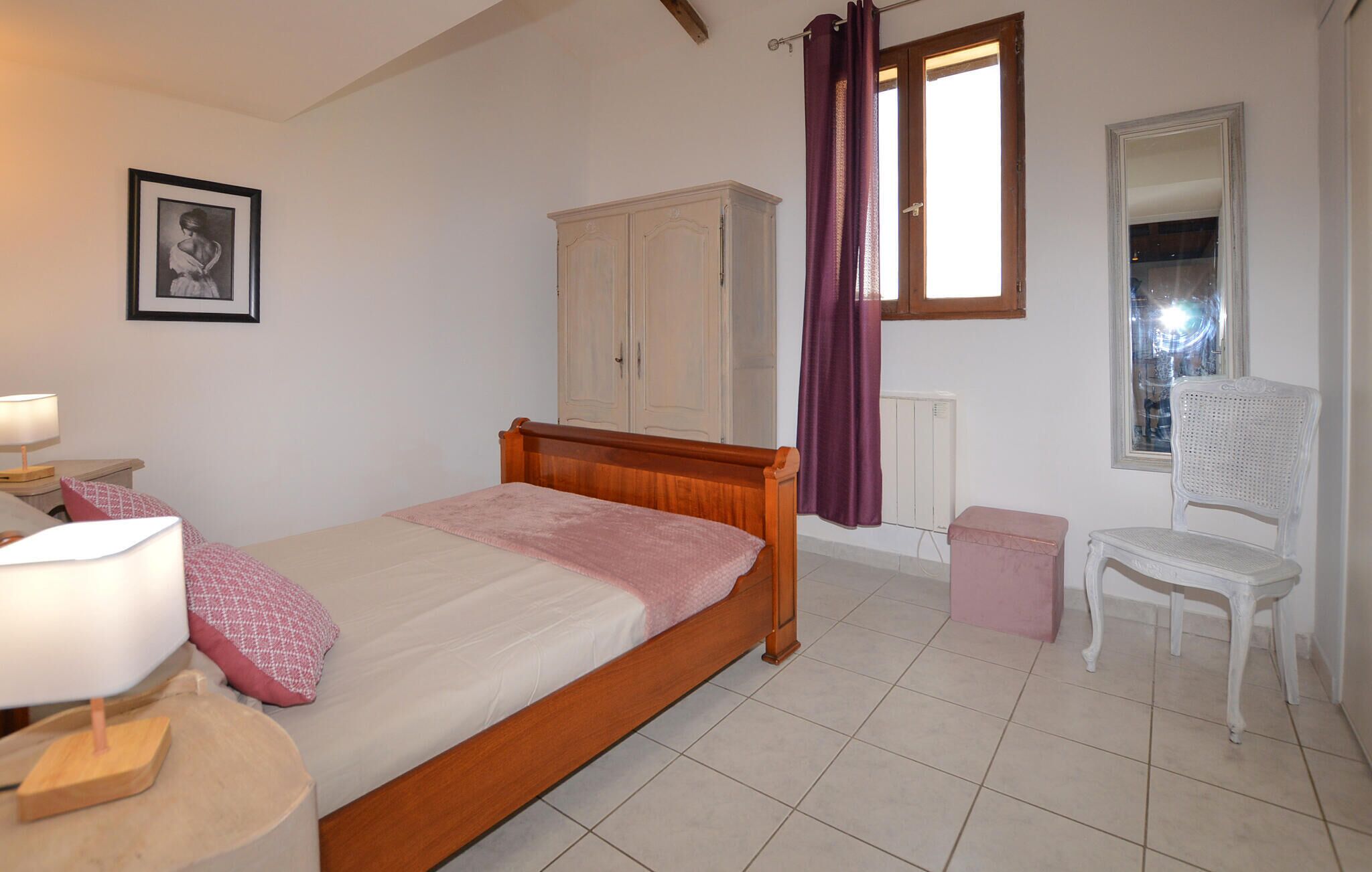 A bedroom with a wooden bed, a nightstand with a lamp, a wardrobe, a window with curtains, a radiator, and a chair.