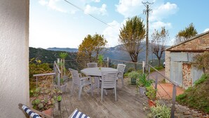 A wooden deck with a round table and chairs, surrounded by potted plants and overlooking a mountainous landscape. - Cozy apartment in Cuttoli with WiFi (Cuttoli)