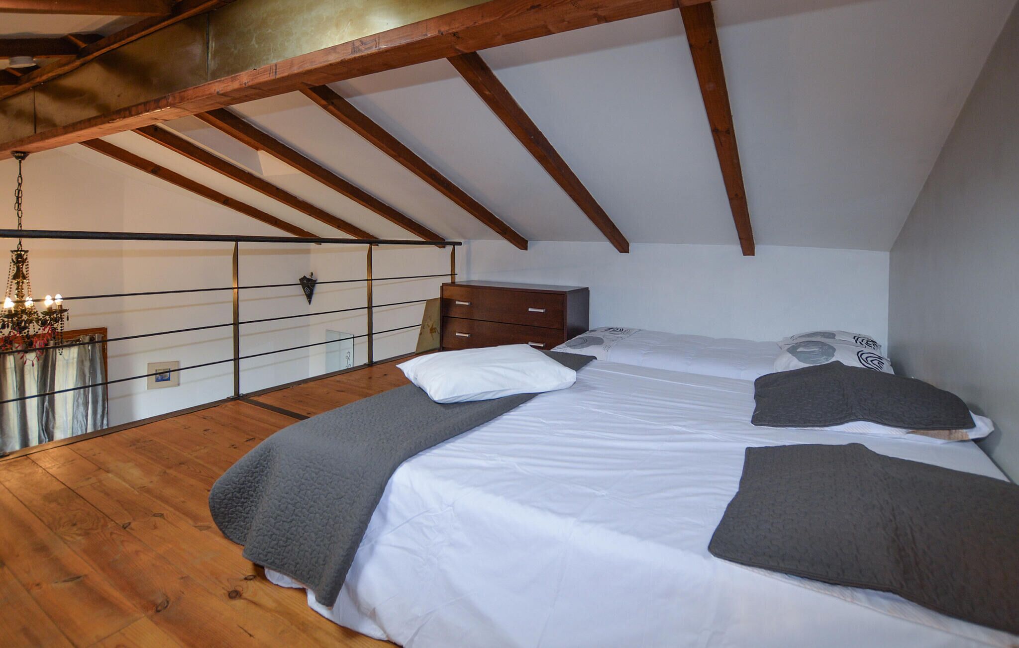 A bedroom with a sloped ceiling, wooden beams, a bed with white and gray bedding, a nightstand, and a dresser.