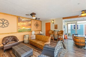 A cozy living room with a brown leather sofa, a cushioned armchair, and a wicker ottoman. There are wooden barrels, a fan, and a ceiling fan. The room has a wooden floor and a painting of a sunset on the wall. - Gulfport Retreat with Wine Cellar Charm (Gulfport)