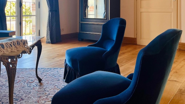 Ein Zimmer mit Holzboden, einem großen Spiegel, zwei blauen Stühlen, einem Tisch mit weißen Spitzen und einem gemusterten Teppich.