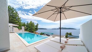 Ein Poolbereich mit Liegestühlen und Sonnenschirm, mit Blick auf ein Gewässer und Grünflächen.