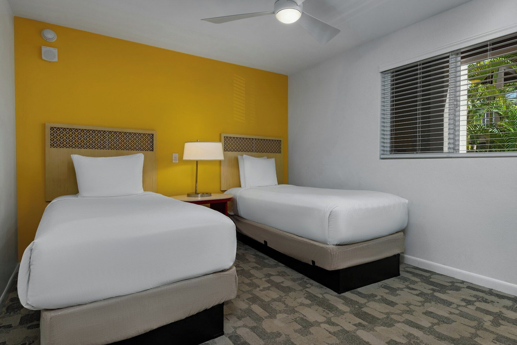 Una habitación de hotel con dos camas, un ventilador de techo, una ventana con persianas y una pequeña mesa con lámpara.