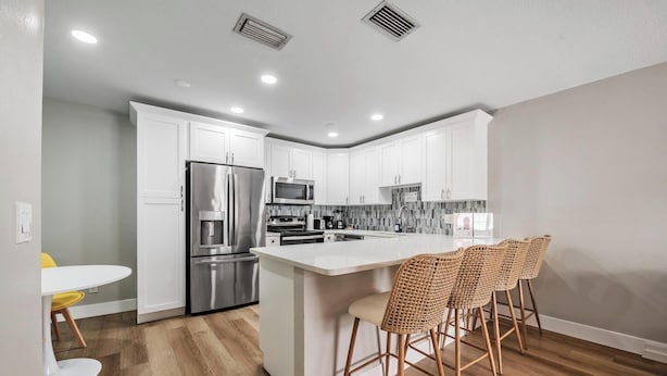Una cocina moderna con armarios blancos, electrodomésticos de acero inoxidable, una isla central con taburetes y suelo de madera.